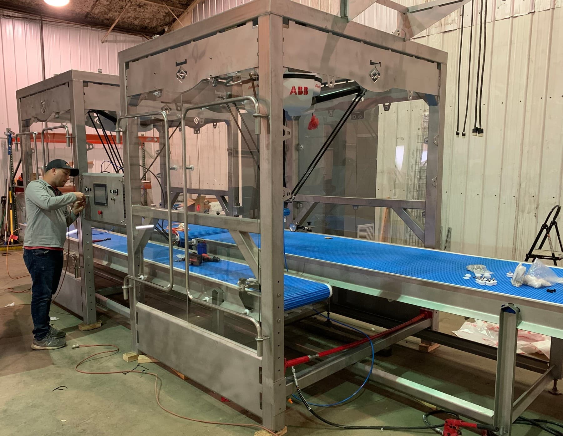 Two stainless robotic pick-and-place cells under construction on the AQS shop floor, each with a delta robot overhead and a blue modular belt conveyor running through the cell, while a technician wires the control panel
