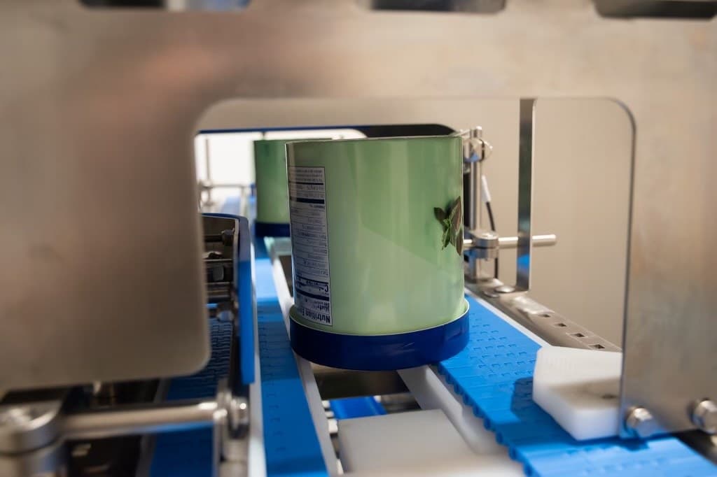 An ice cream tub riding two independent blue belts inside the stainless inspection tunnel