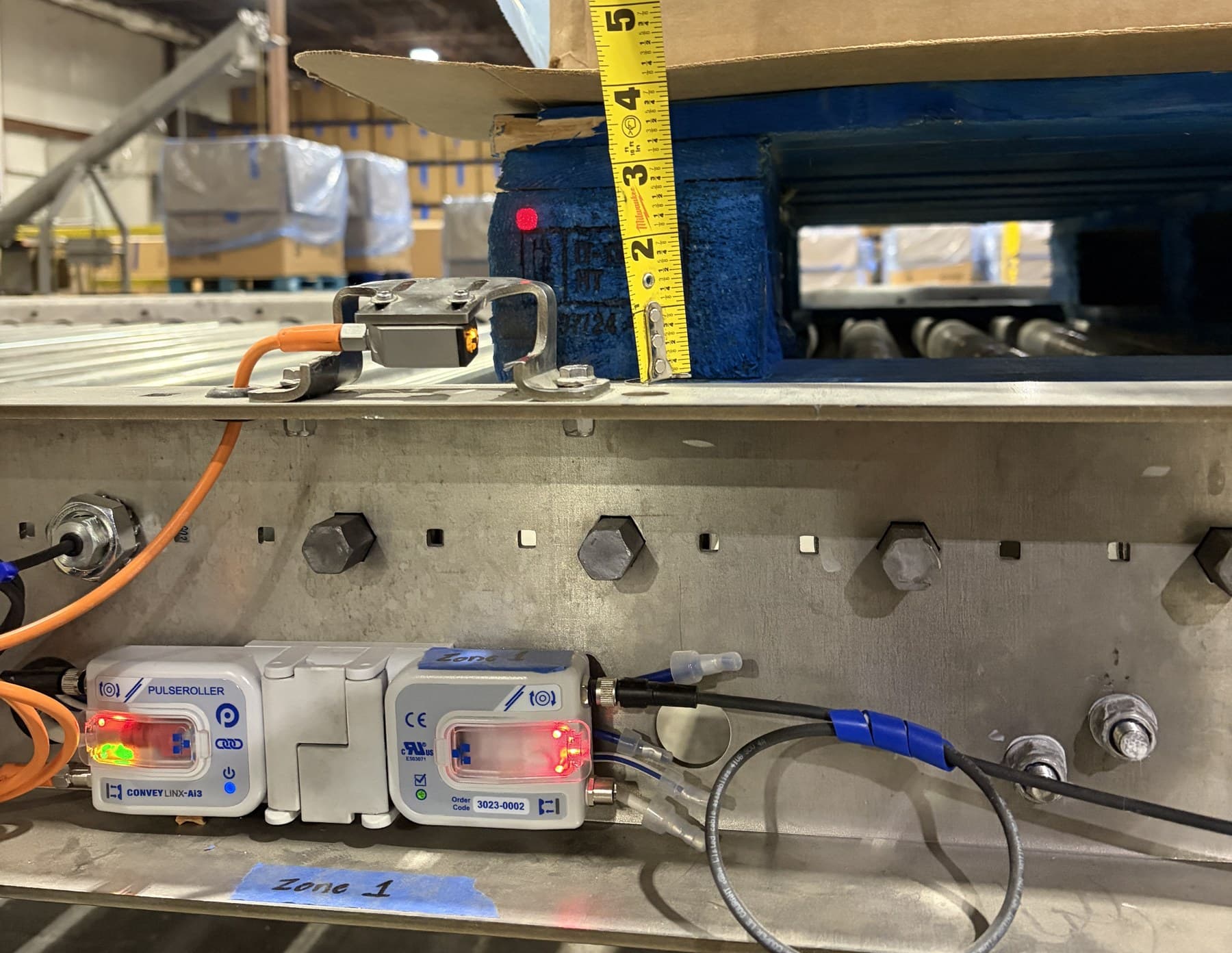 A diffuse photo eye on a stainless MDR conveyor frame aimed at the painted stringer of a blue pallet, with the zone's 24 VDC motor-roller card mounted below